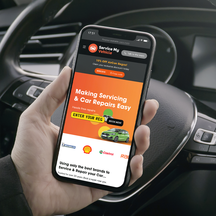 Person holding a smartphone inside a car, displaying the 'Service My Vehicle' website. The screen shows a promotional offer for 10% off air conditioning repairs, a call to action for easy car servicing and repairs, and logos of trusted brands like Michelin, Shell, Castrol, and RAC. The background shows the car's steering wheel and dashboard.