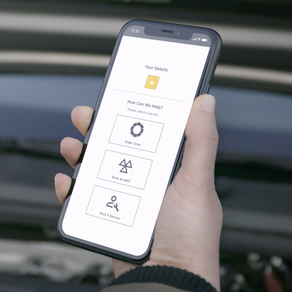 Person holding a smartphone displaying a vehicle service app screen. The app shows options to order tyres, book an MOT, and book a service under a section titled 'How Can We Help?' with a simple, clean interface. The background shows the car's open hood.