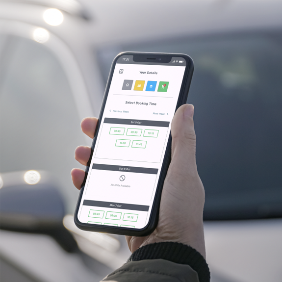 Person holding a smartphone displaying a vehicle service app screen for selecting a booking time. The interface shows available time slots for different days of the week. The background features a blurred view of a parked car.