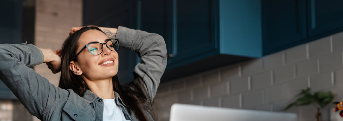 A woman wearing glasses and a grey shirt leans back with her hands behind her head, smiling and appearing relaxed. She is sitting in a kitchen environment with blue cabinets and white tile backsplash in the background.